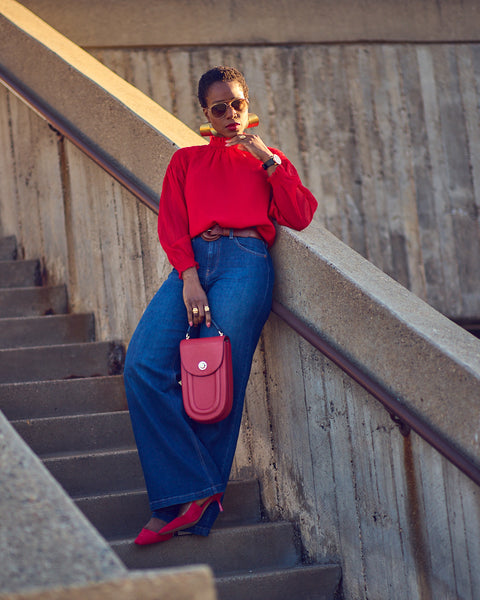 Style influencer Farotelle wearing a red and blue color-blocked outfit. She has on blue wide-leg jeans with a bright red blouse, red pumps, and a red oval-shaped handbag. She's standing on stair steps and is wearing sunglasses and oversized gold earrings.