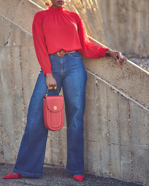 Style influencer Farotelle wearing a red and blue color-blocked outfit. She has on blue wide-leg jeans with a bright red blouse, red pumps, and a red oval-shaped handbag. She's standing next to a wall and is wearing oversized gold earrings.