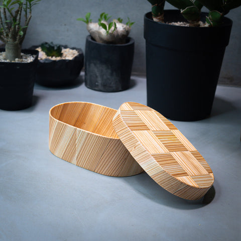 A wooden box with a herringbone pattern on the lid rests on a concrete surface. In the background, there are three potted plants of varying sizes and colors, creating a contrast with the natural wood and the industrial feel of the grey background.