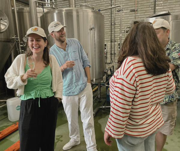 4 people standing in a brewery, holding glasses of prosecco in a celebration, laughing and wearing fauna branded hats.
