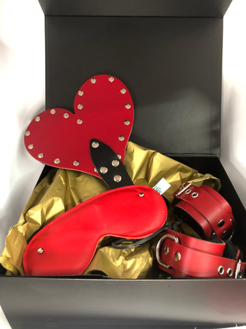 A black gift box with gold tissue paper. Inside box: A red heart-shaped paddle, a red blindfold, red cuffs and a red collar.