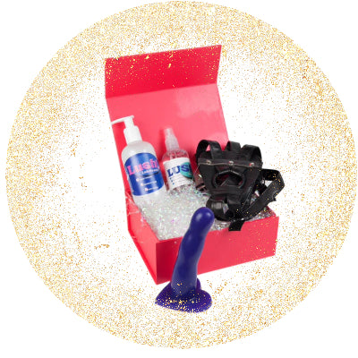 A red gift box with a bottle of lube, a bottle of toy cleaner and a black leather strap-on harness. A purple dildo is in front of the box.