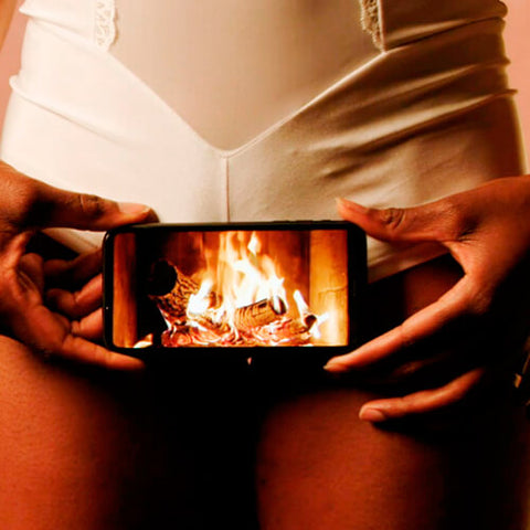 A WoC in white lingerie holdiong a mobile phone against her genitals. On the screen is a fire burning. 