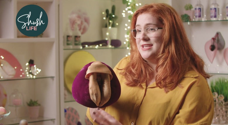 A woman with long red hair, glasses are dark yellow shirt holding a plush vulva puppet.