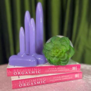 Deep green curtains background. Pale pink table cloth. our lilac vaginal dilators on top of two copies of Becoming Orgasmic. A small green succulent for decoration. 