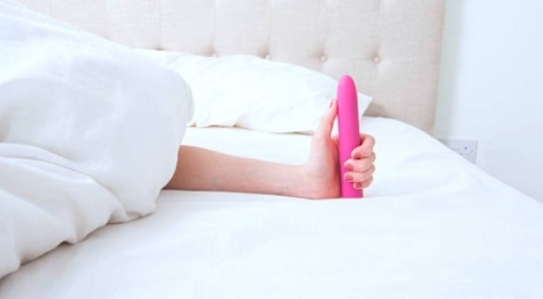 white bedding. a woman's arm is stretched out from under the covers, and she's hliding a bright pibk classic-style vibrator