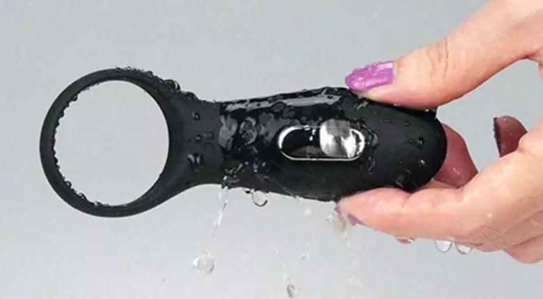 A hand with purple nail varnish holding a black cock ring. Water is splashing.