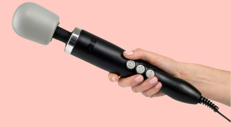 Blush pink background. A white hand with no nailvarnish holding a Doxy wand massager in black and grey,