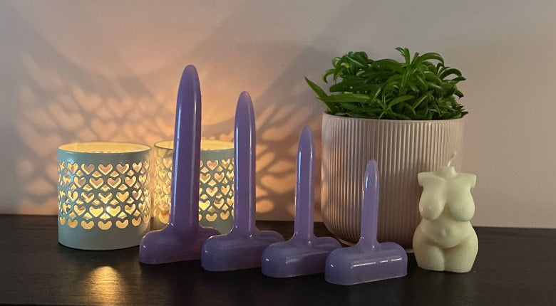 Four lilac vaginal dilators in progressive size. Backgropund: 2 white candle holders, a green plant in pa;e pink bioth ana white candle in the shape of a curvy woman