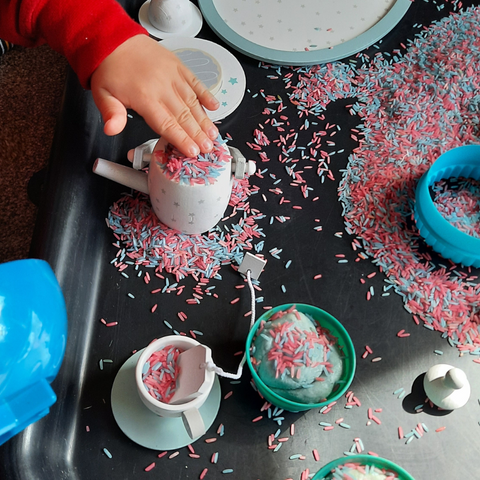 Toddler's hand placing a lid on a tea pot full of blue and pink sensory rice