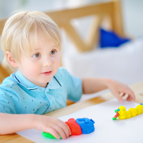 Male toddler with blonde hair sitting at a table making a colourful caterpillar out of clay
