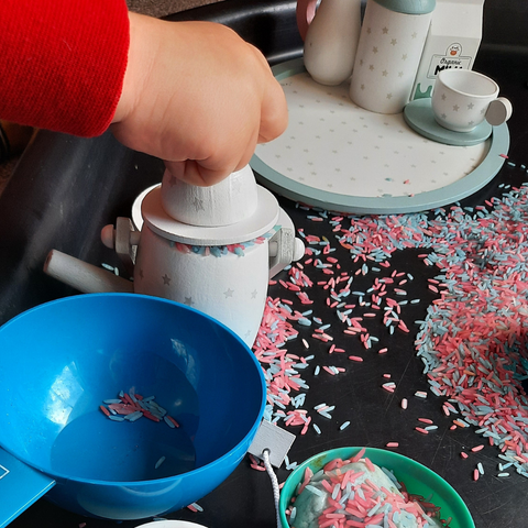 Toddler's hand sprinkling sensory rice into a wooden tea pot on the tea party messy play tray