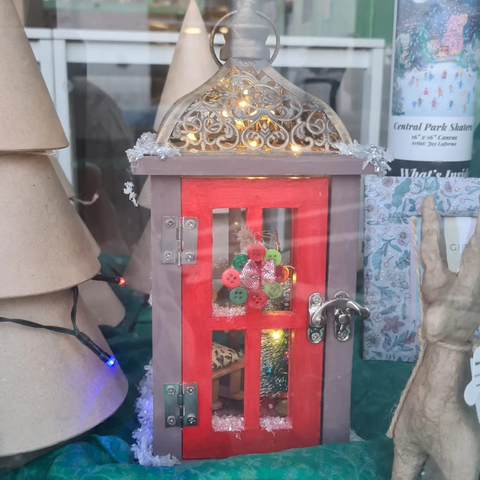 Grey Christmas lantern with a red door covered in snow and sparkly gold glitter