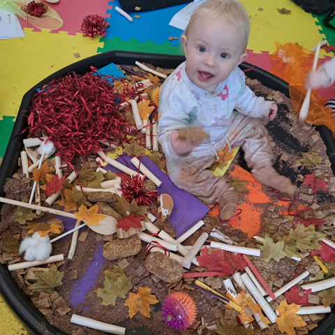 Image shows a happy baby sitting on a tuff tray surrounded by red tissue paper, wooden spoons, cereal, craft leaves and wooden hedgehogs