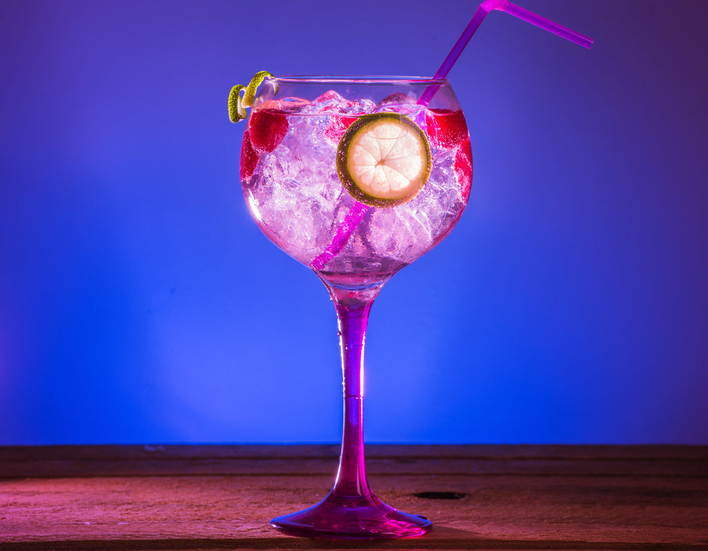 Taça de Pink Gin com gelo, um dos tipos de gin, sobre uma mesa de madeira e um fundo azul