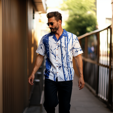 A man with styled facial hair walks along a narrow pathway next to a wooden building. He is wearing a short-sleeved white shirt with a blue bamboo pattern, paired with dark pants. His posture is relaxed and confident as he strides forward.