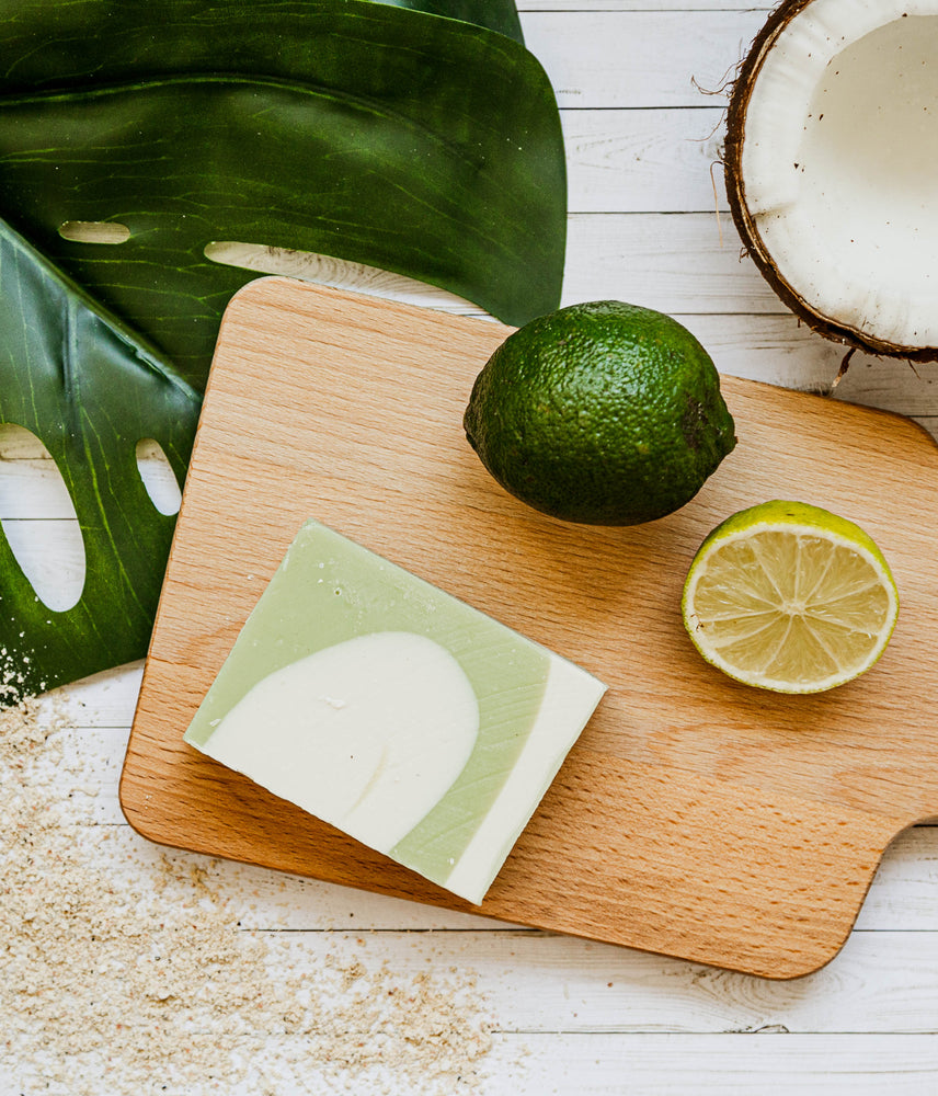 A green and white swirled bar of soap rests on a wooden cutting board with a cut lime, a whole lime, and an open coconut and big leaf in the background. There is sand scattered across the table top.