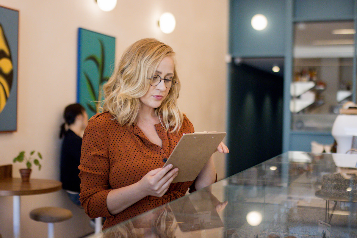 Blonde woman wearing round Manhattan Reading Glasses reading from a clipboard at a table.