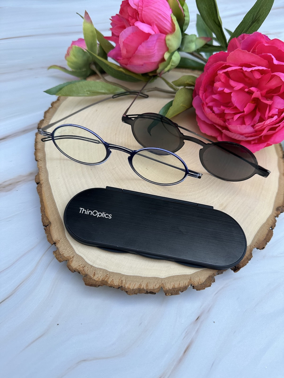 A pair of round Manhattan Reading Glasses and round Manhattan Reading Sunglasses next to each other on a wooden plate alongside a Milano Case and flowers