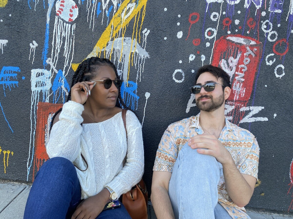 Man and woman sitting on a sidewalk wearing ThinOptics Wanderlust Sunglasses while looking at each other.