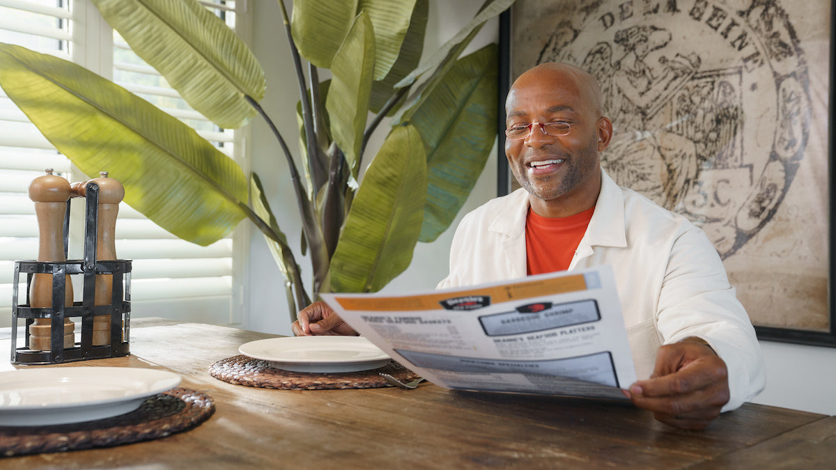 Smiling man reading a menu at a restaurant with ThinOptics reading glasses perched on his nose, exemplifying the practical elegance of the eyewear. The scene highlighting how ThinOptics glasses and Readers fit seamlessly into daily life.