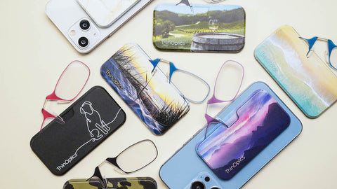 Array of ThinOptics readers with their printed Universal Pod cases featuring vibrant designs such as landscapes, abstract art, and a dog silhouette, displayed alongside smartphones, demonstrating the stylish customization options for carrying ultra-portable reading glasses.