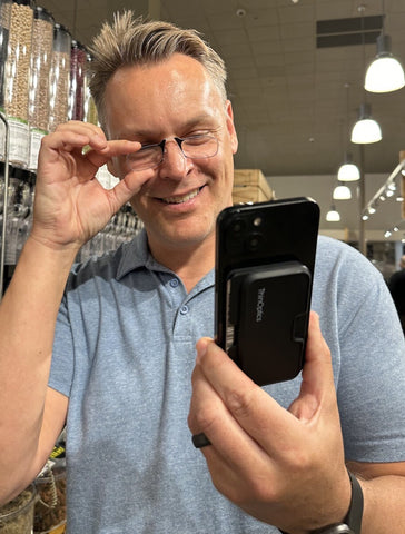 A smiling man wearing Reading Glasses and holding a phone with an attached ThinOptics Universal Pod Wallet in his other hand, in a grocery store aisle.