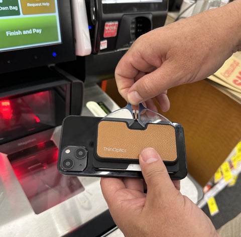 A person pulling out Readers from their ThinOptics Universal Pod Wallet in brown faux leather attached to the back of a smartphone with MagSafe® technology at a grocery store checkout stand.