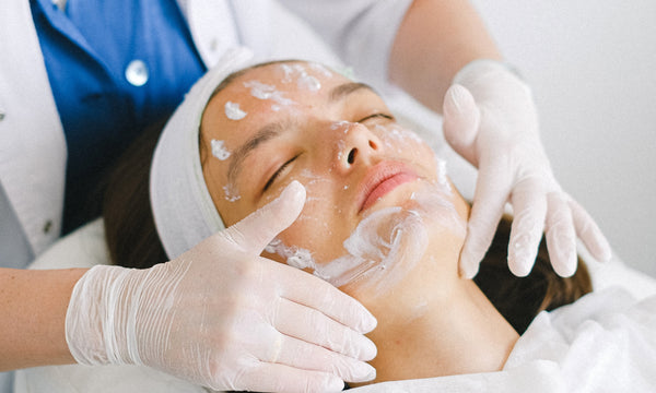 woman with her eyes closed getting cream spread on her face by a clinician wearing white gloves