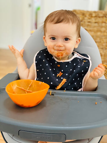 Baby girl with black and white bib and an orange bowl full of spaghetti