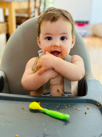 Baby hugging a jar of baby food with green spoon in high chair