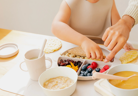 Baby exploring berries and other food in plate with mom's hand helping