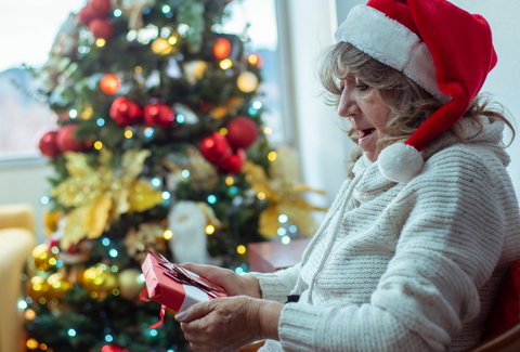 Grandma in white sweater and Santa hat in front of Christmas tree opening a present