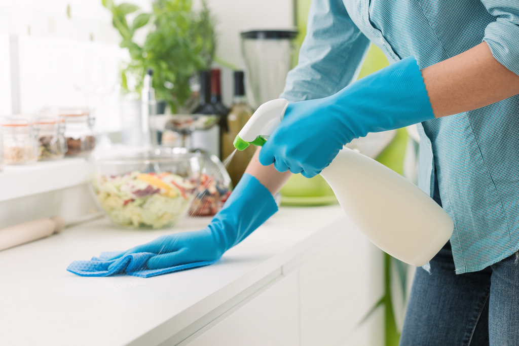 A woman wearing blue rubber gloves cleans a counter top with a spray bottle and cloth. 