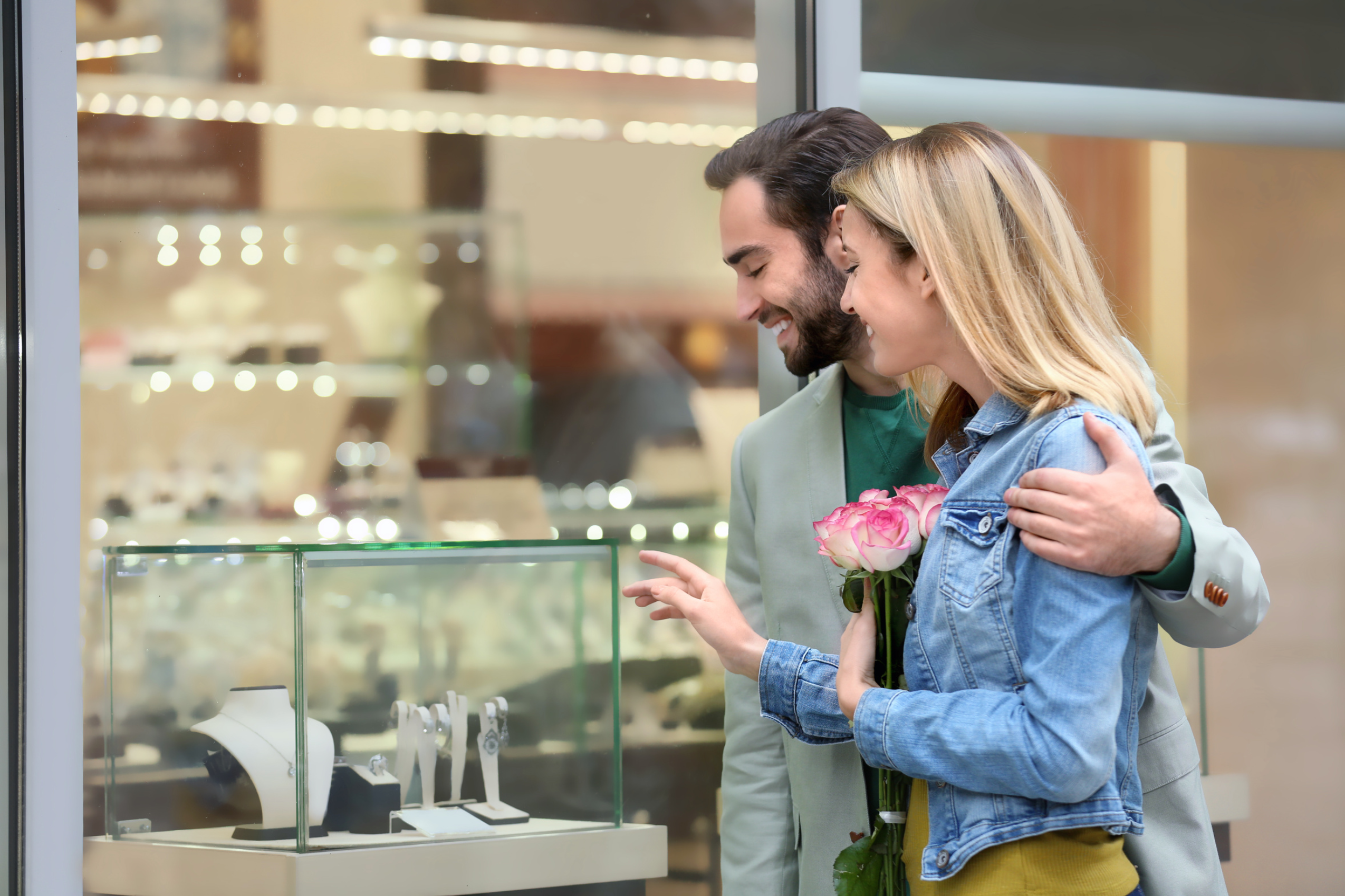 A man and woman look in a glass case at a jewelry store while shopping for bridal rings. The woma holds a bouquet of roses. 