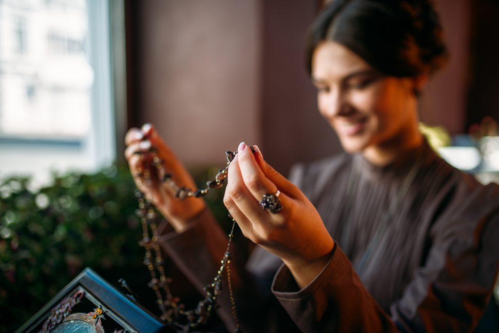 A smiling woman holds an antique necklace as she decides whether to put it on. 