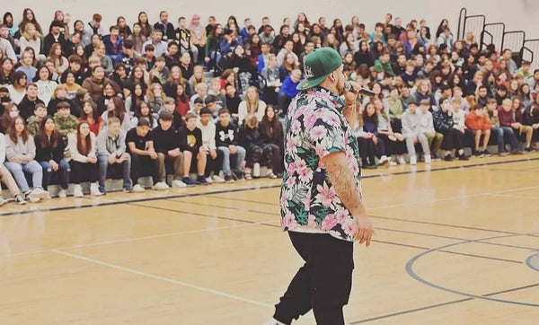 Coaster Speaking at a local highschool. He's has a mic in his hand and is talking to a crown of teenagers in the gymnasium.