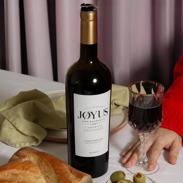 A bottle and glass of Joyus Non Alcoholic Cabernet Sauvignon on a table with sliced bread and olives