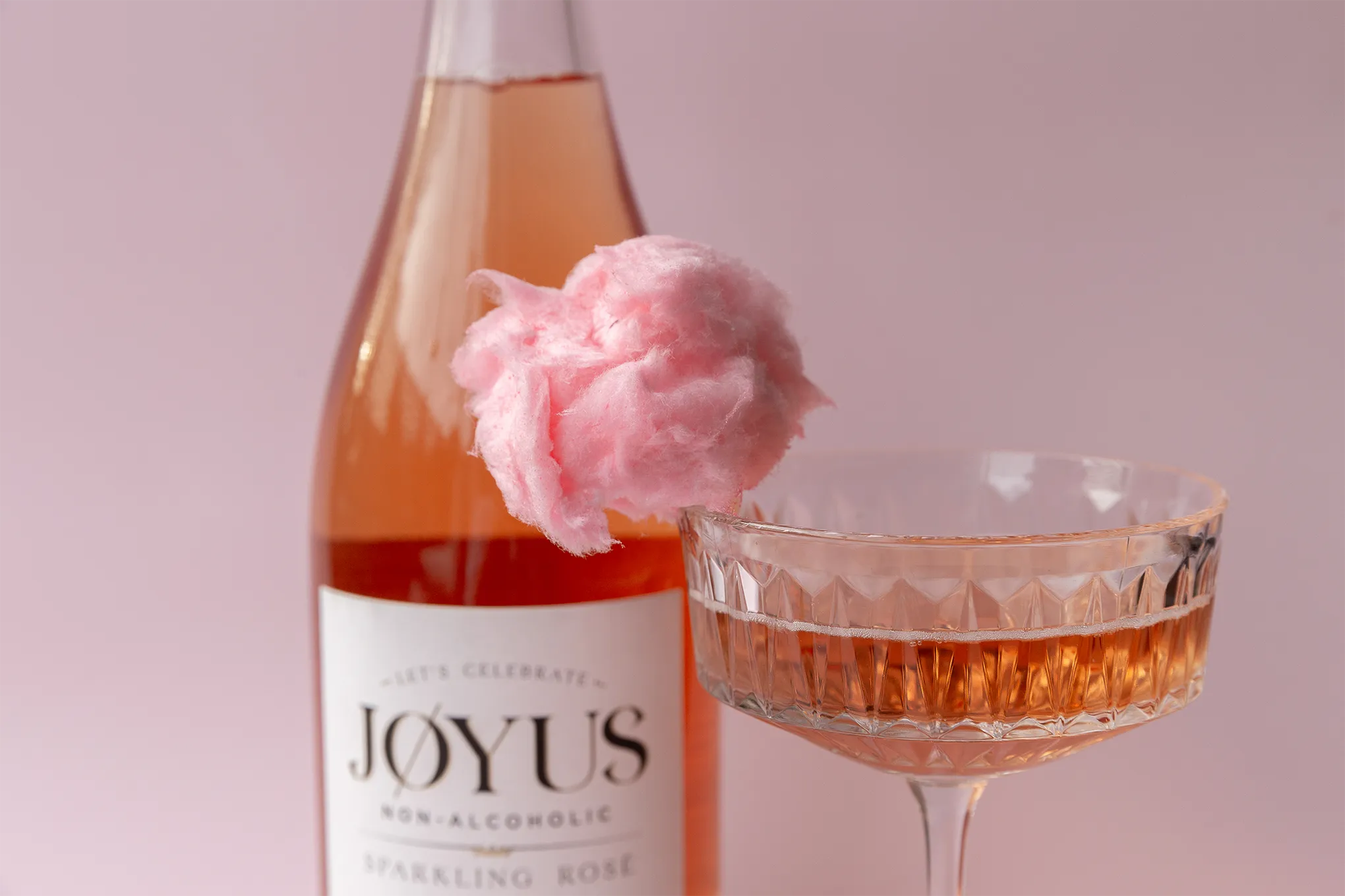one bottle of Joyus nonalcoholic sparkling rosé close up. Also in frame is a close up of the fluffy pink cotton candy garnish on a cut crystal coupe champagne glass with Joyus in it.