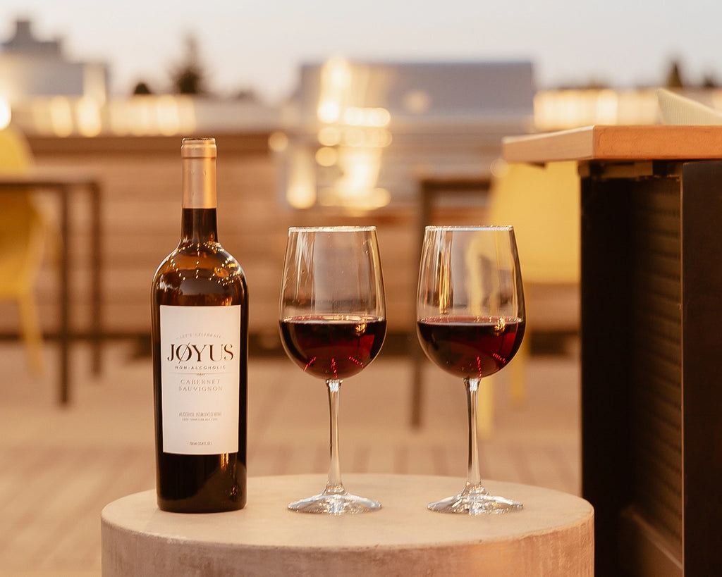 A bottle and two glasses of Joyus Cabernet Sauvignon in front of an urban skyline at dusk.
