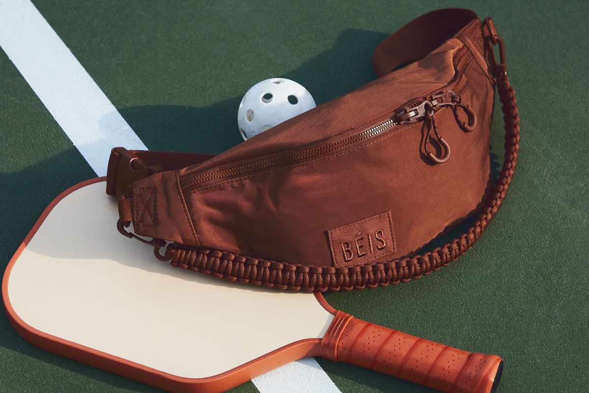 Brown crossbody sling on top of a pickle ball paddle next to a whiffle ball