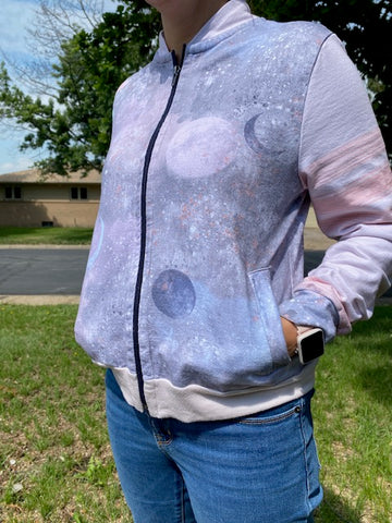 A bomber jacket with light pink sleeves with darker pink stripes. The body of the jacket is has a modeled gray background with light pink and blue moons and planets overlayed.