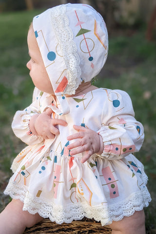 A baby wearing an off white dress and bonnet with 1950's geometric shapes in orange, blue and green on it.