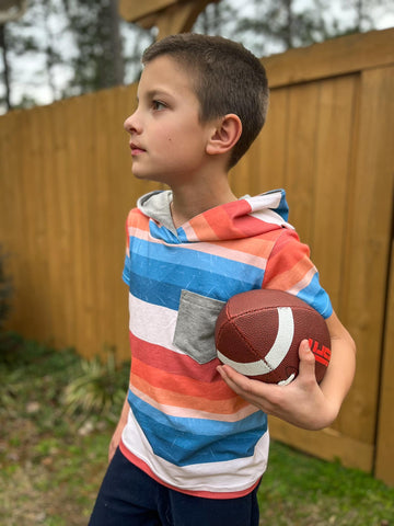 A little boy with a football wearing a hooded T-shirt with shades of orange and shades of blue stripes.