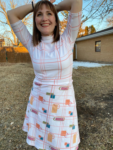Girl standing outside wearing a turtleneck shirt with lines on it and a skirt with orange and green boxes on it.