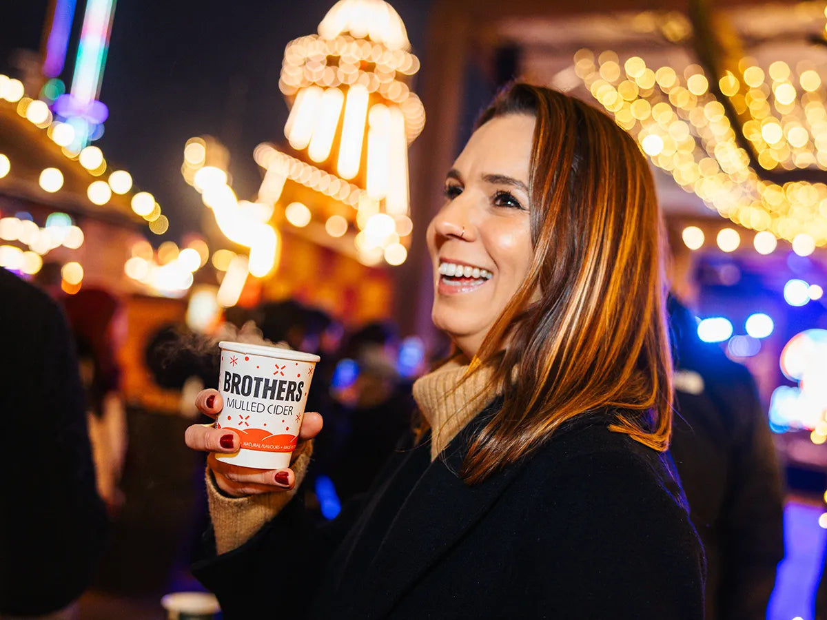 Woman holding a cup with 'Brothers Mulled Cider' at a festive outdoor event with lights.