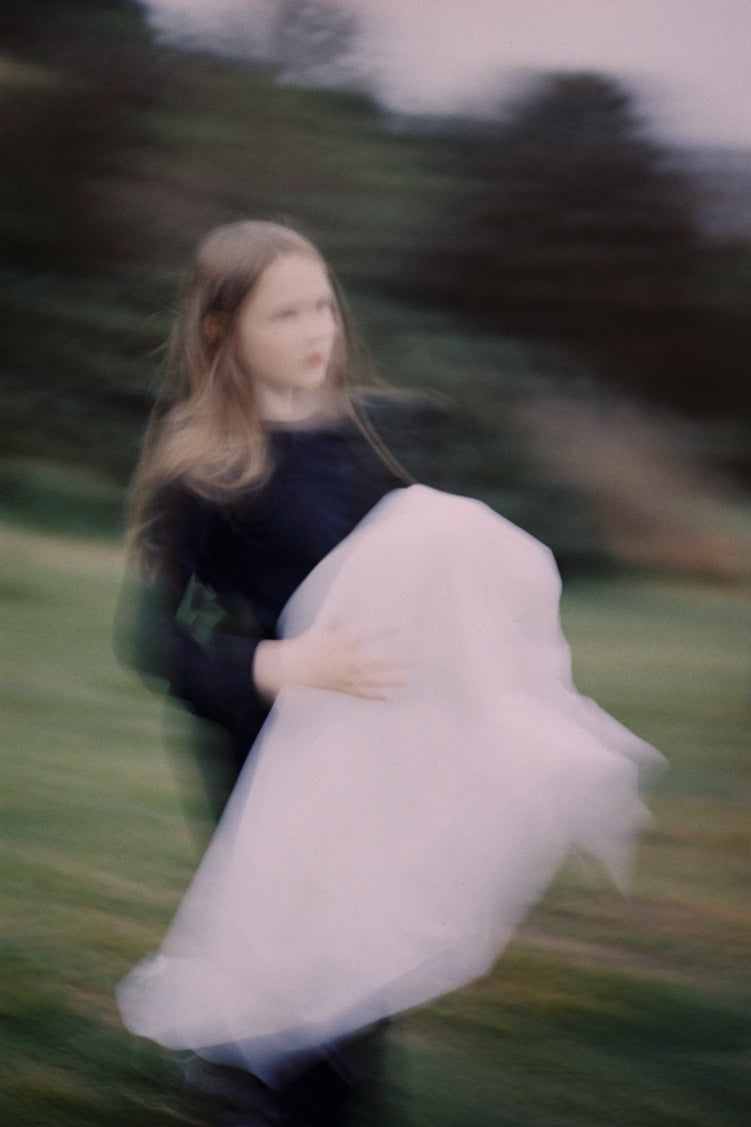  A photograph taken at the Wings workshop. Photo shows a young girl holding a bundle of white fabric. The image is blurred, and in the background are bushes, grass, and trees.