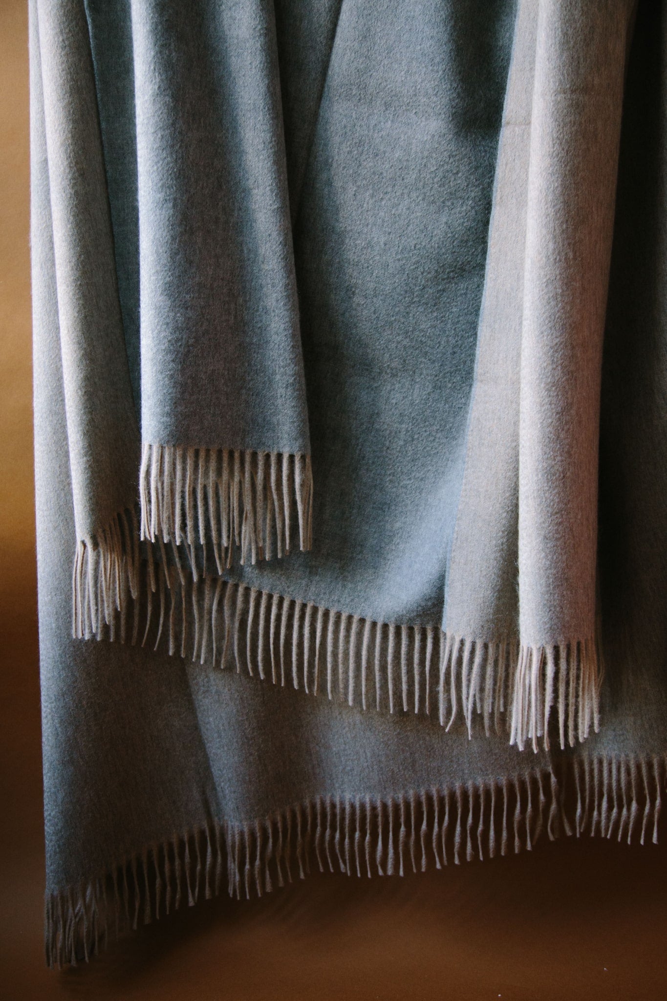 Large cashmere throw. Two sided reversible colour; grey and pale beige. Draped against a brown background with natural light.