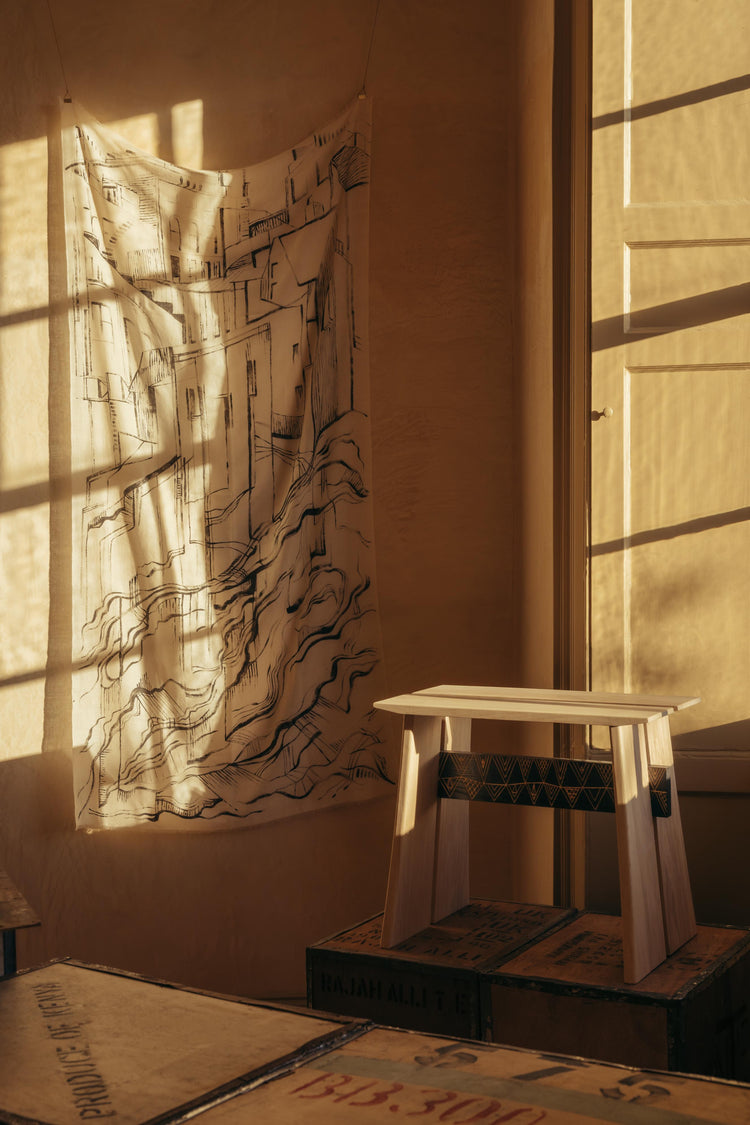  The Brodgar Stool by The Orkney Furniture Maker. Light from the window to the right of the stool casts warm sunlight across it's top. The stool is made from Ash and has a charred, patterned panel beneath the seat.