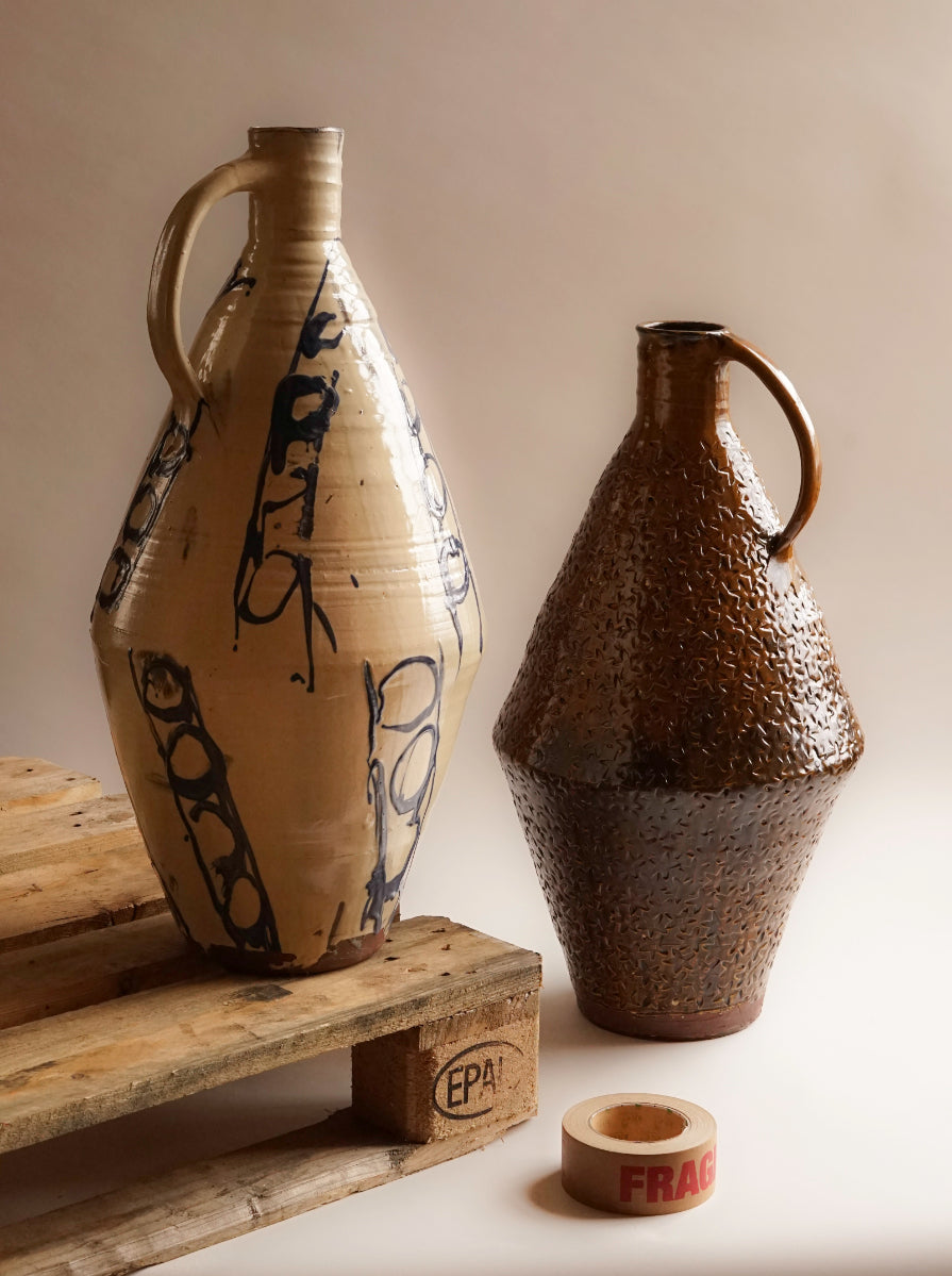  Two large ceramic flagons. With curved handles connected to the mouth of the vessel. The cream flagon on the left of the image has expressive circular markings. The flagon on the right, is a deep amber colour, and has been pressed with a stamp all over it's surface.